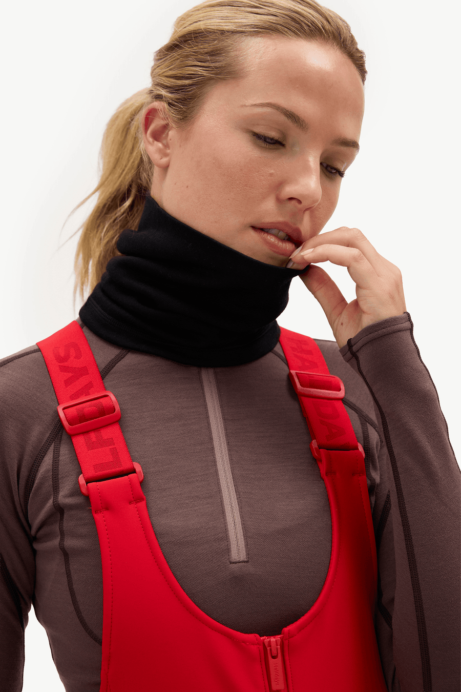A close-up of a woman wearing a black neck gaiter and a red bib over a brown long-sleeve athletic top, against a plain light background.
