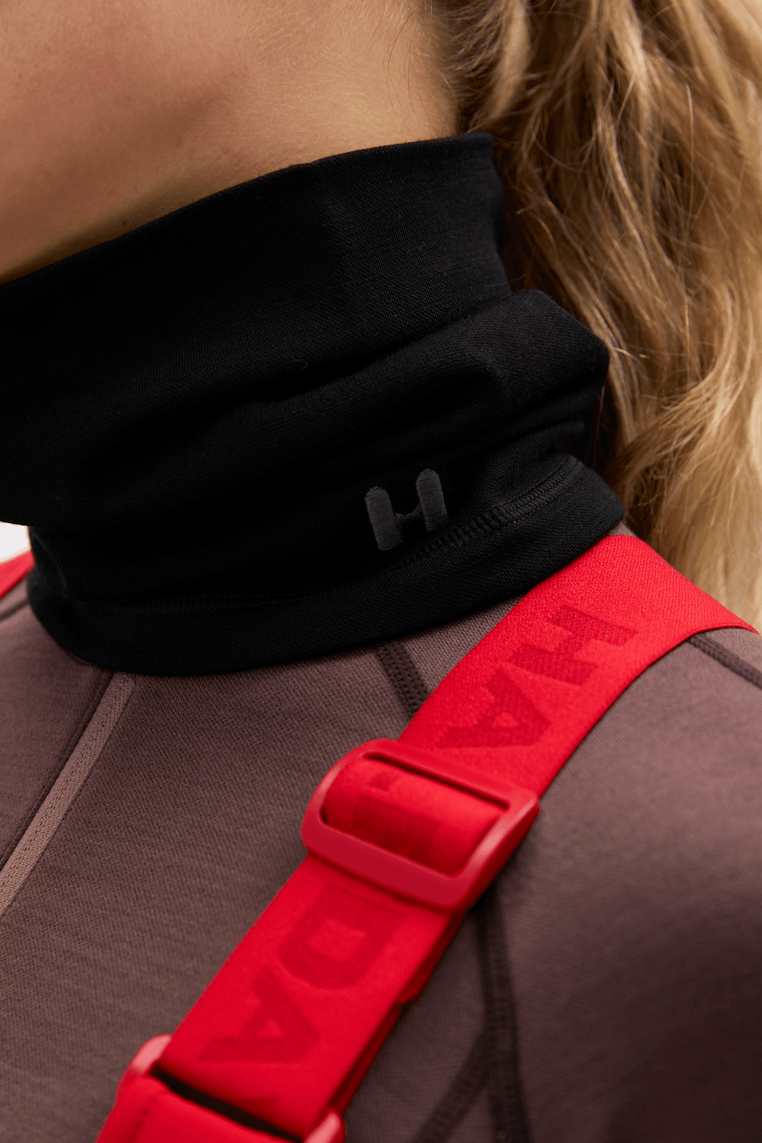 Close-up of a person wearing a black turtleneck-style neck gaiter with a small embossed H logo, paired with a brown athletic top and a bright red shoulder strap with embossed lettering.