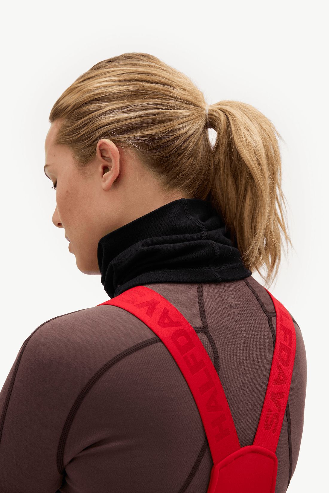 A woman shown from the back and side wearing a dark brown athletic wetsuit with black seam lines, a black neck gaiter, and bright red shoulder straps with the brand name HALIDAY printed in red.