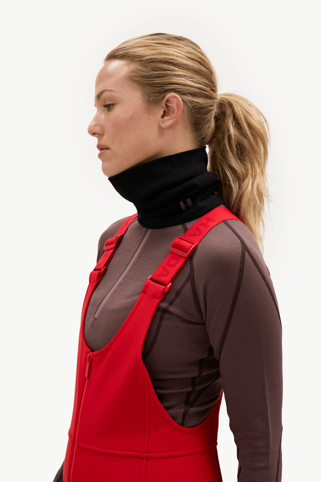 A woman in profile wearing a black neck gaiter and red bib overalls over a brown long-sleeve athletic top, photographed against a clean white studio background.