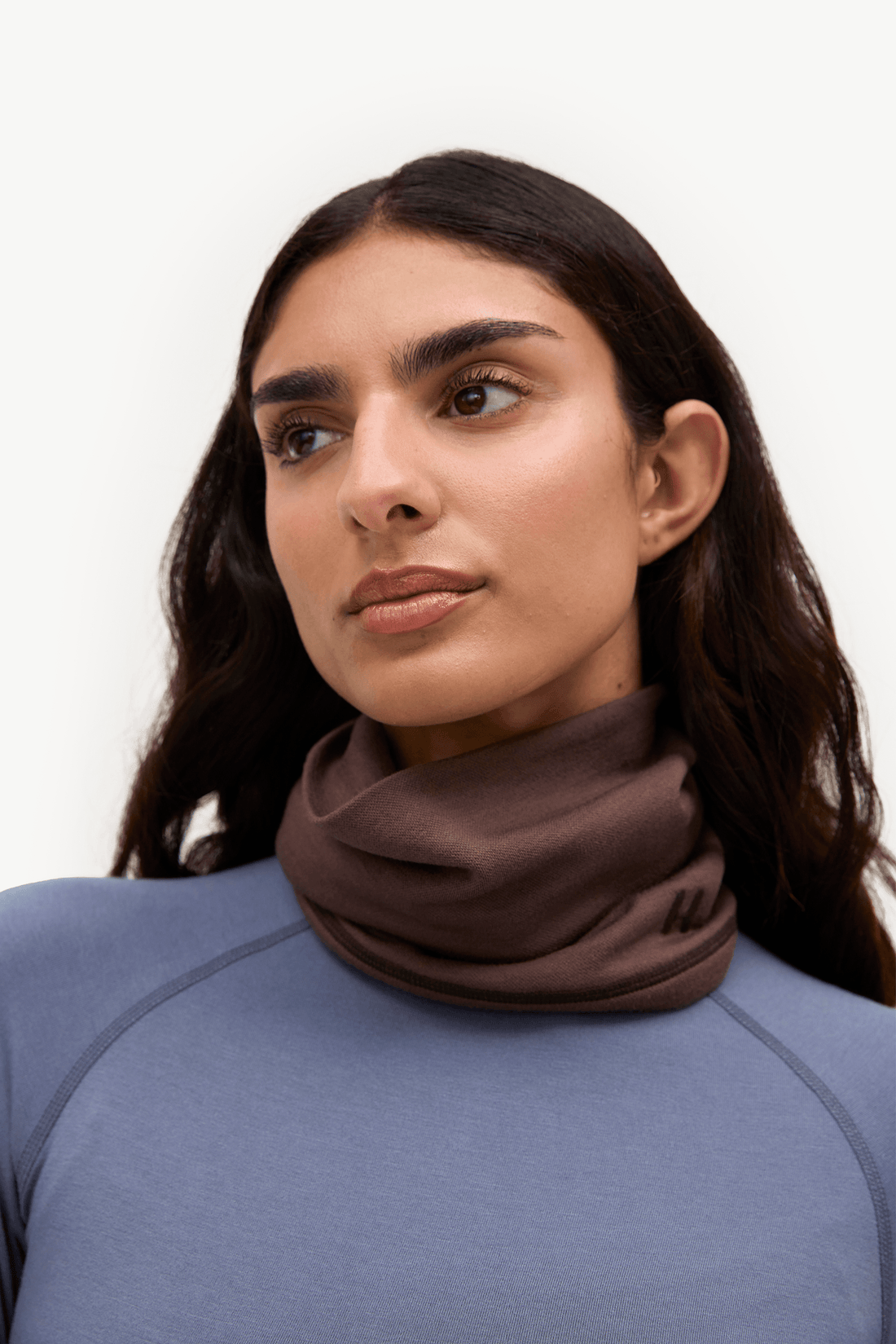 A close-up of a woman wearing a dark brown cowl neck scarf and a slate blue athletic top against a plain white background.