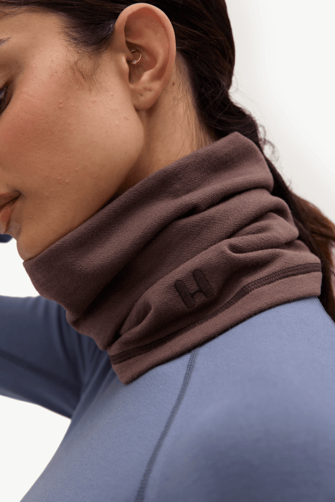 Close-up of a woman wearing a dark brown athletic neck gaiter with a small embroidered logo, paired with a blue long-sleeve top against a plain white background.