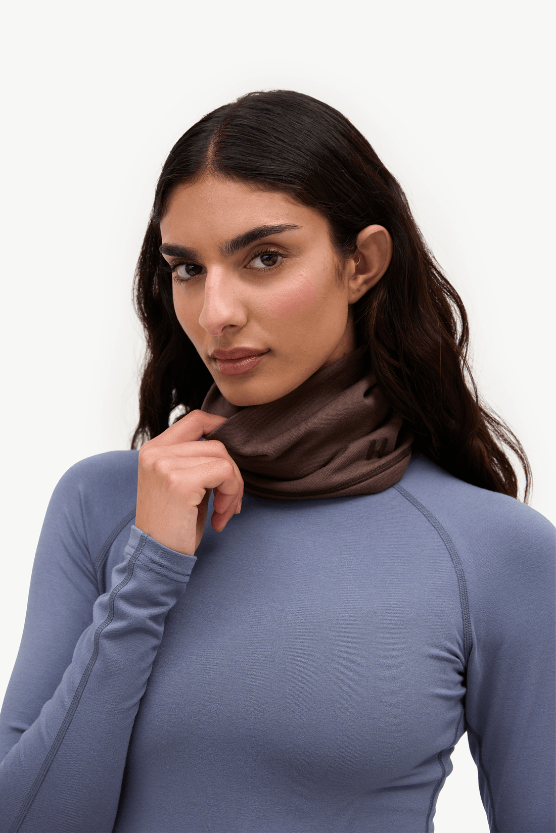 A woman wearing a dark brown athletic neck gaiter and a slate blue long-sleeve top, posing against a clean white background.
