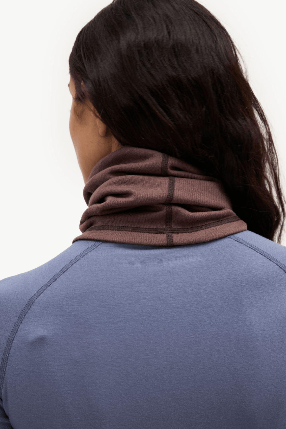 A close-up back view of a person wearing a dark brown athletic neck gaiter over a slate blue-gray long-sleeve top, against a plain white background.