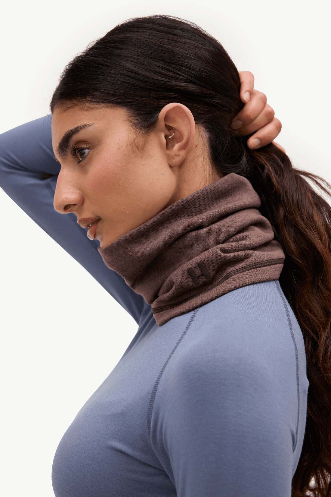 A woman in profile wearing a brown ribbed neck warmer or cowl neck gaiter over a slate blue long-sleeve top, against a plain white background.