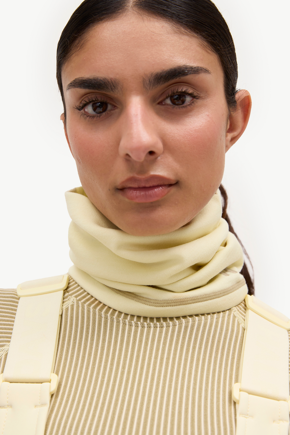 Close-up of a model wearing a pale yellow turtleneck sweater with a matching cream harness or vest over a beige striped top, against a plain white background.