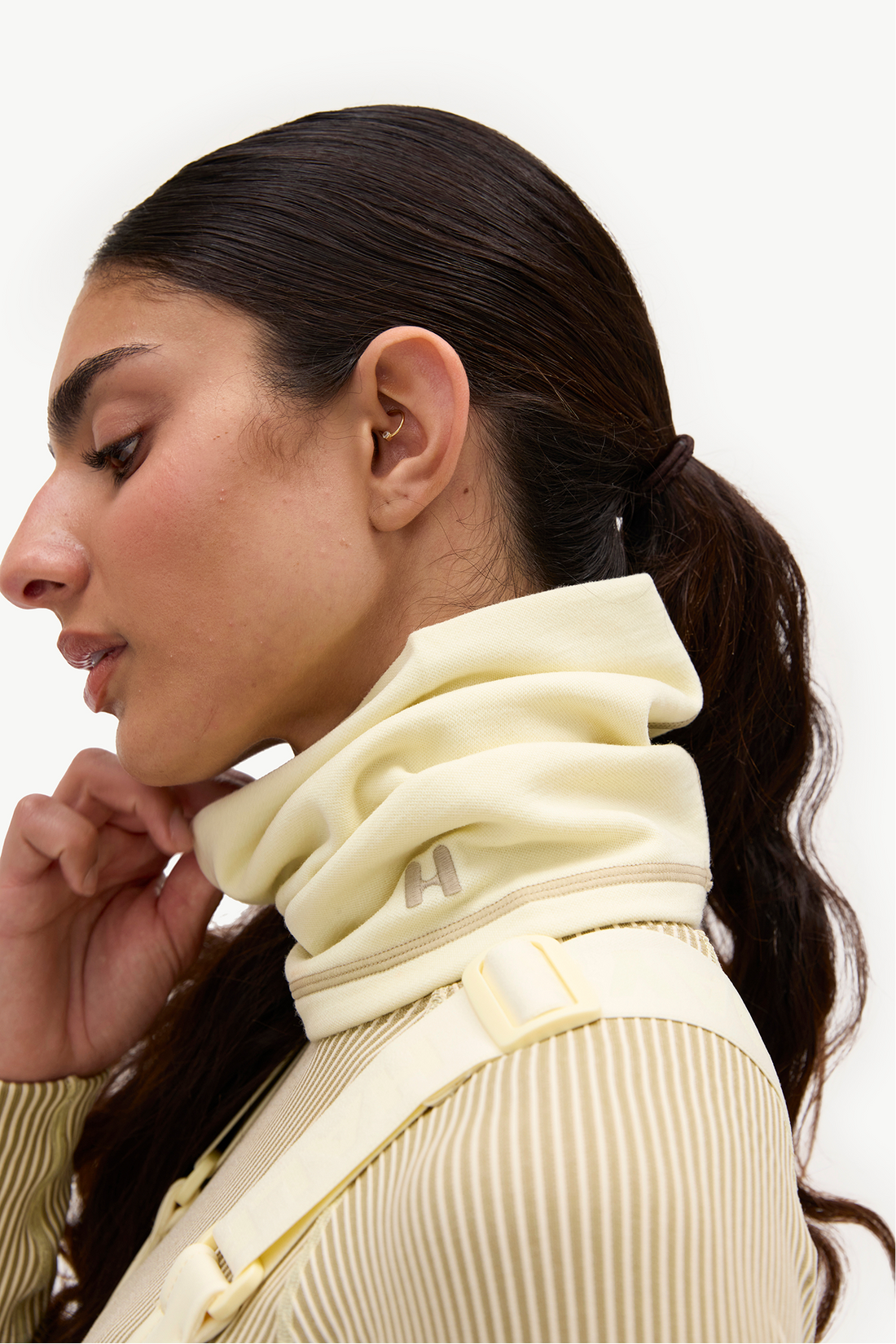 Close-up side profile of a woman wearing a cream-colored neck warmer with a buckle strap and a matching ribbed top against a plain white background.