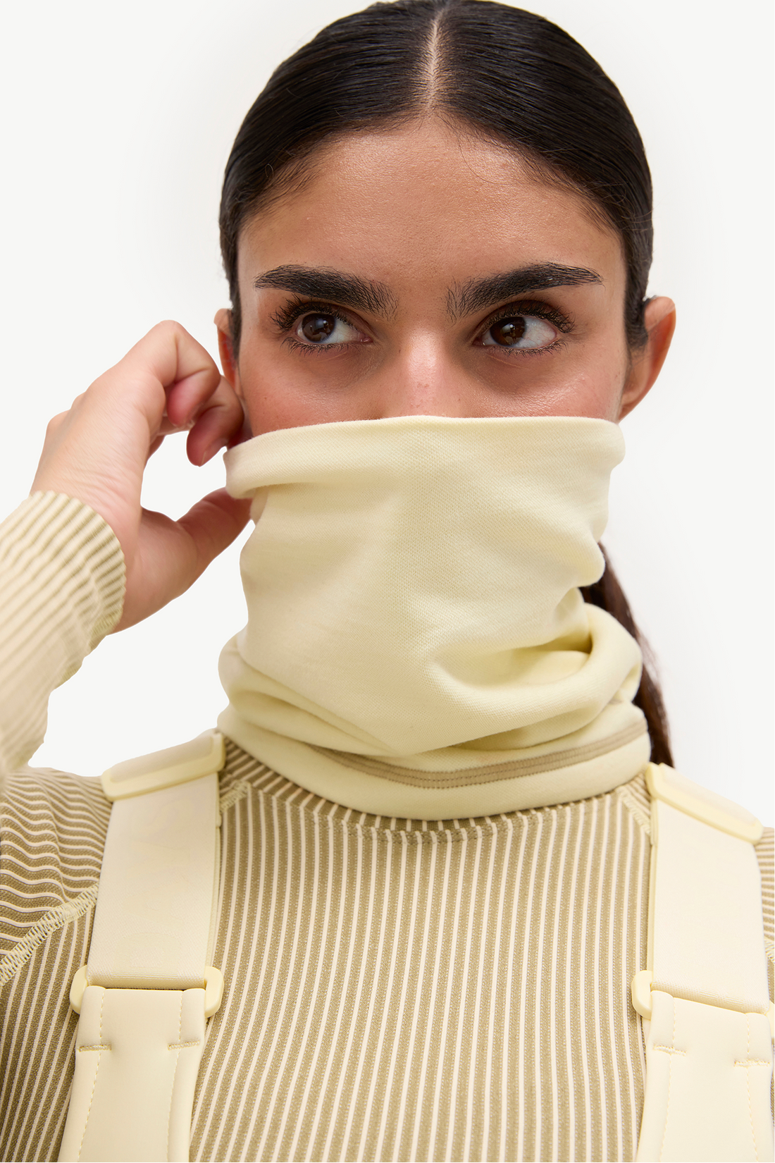 A close-up of a woman wearing a pale yellow fleece neck gaiter or balaclava, pulled up over her nose and mouth, paired with a beige ribbed turtleneck sweater and a cream-colored backpack against a white studio background.