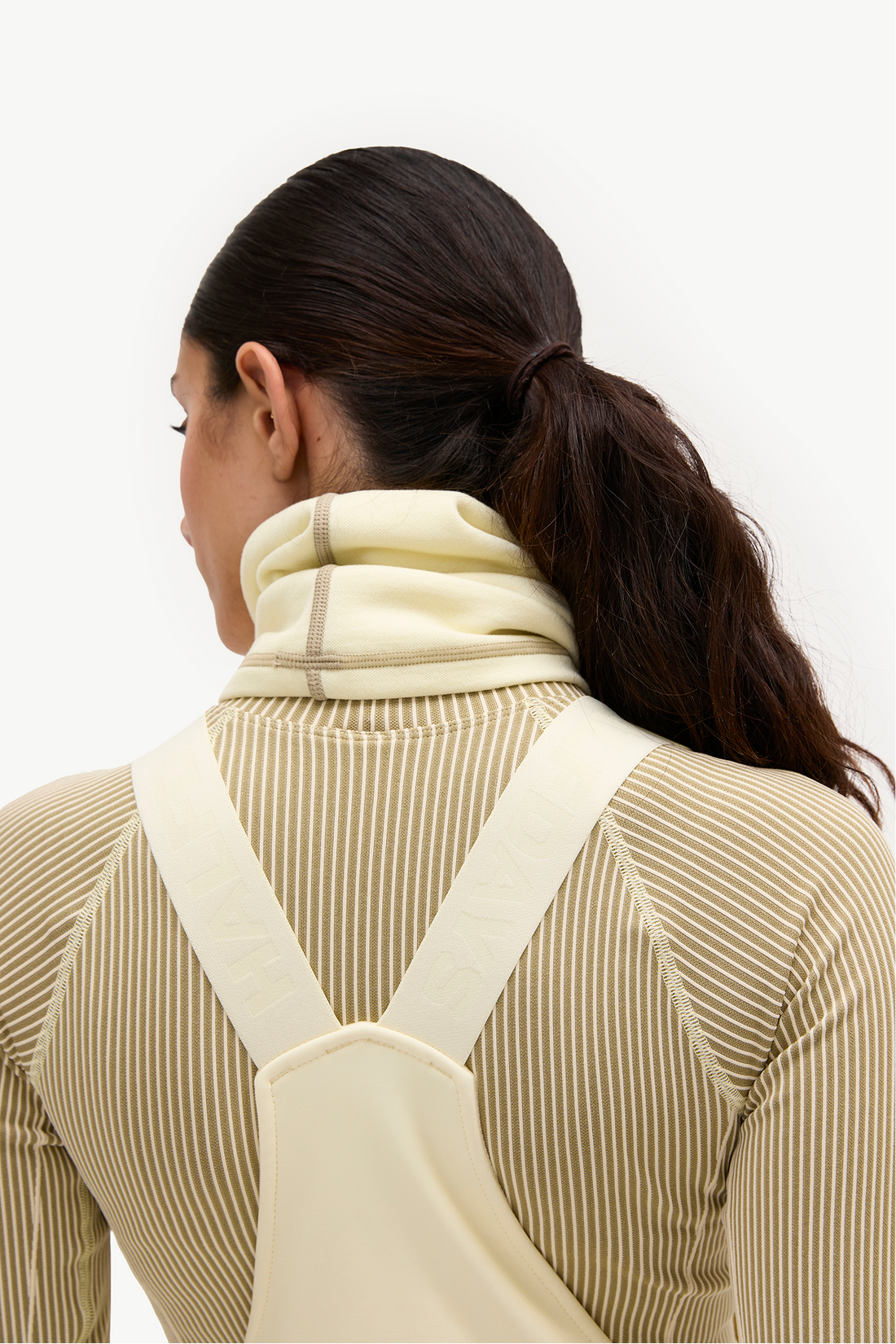 A woman shown from the back wearing a cream-colored undergarment with wide shoulder straps over a beige ribbed long-sleeve top, against a plain white background.