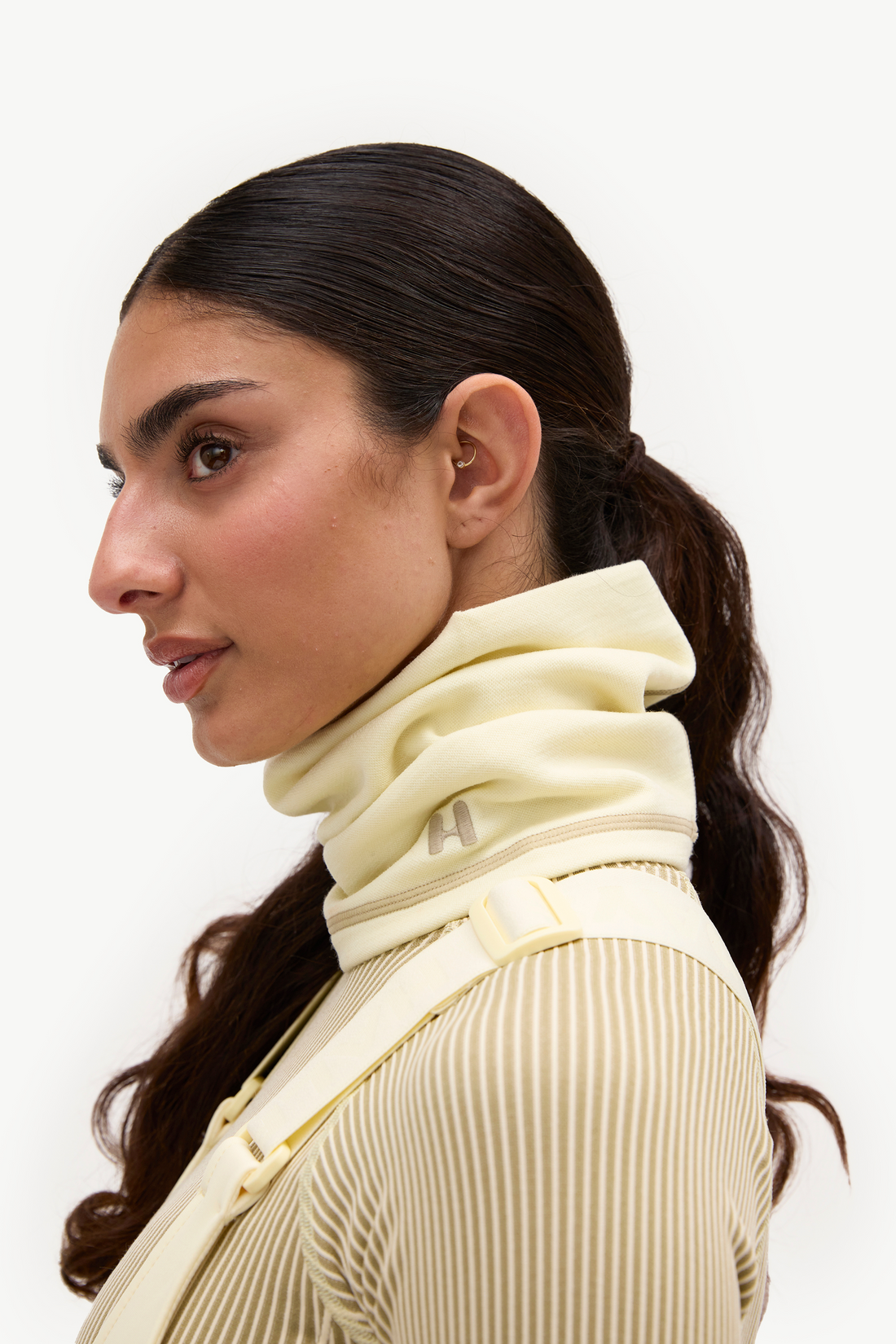 A close-up side profile of a woman wearing a cream-colored ribbed knit top with a matching high-neck collar and shoulder strap, set against a plain white background.