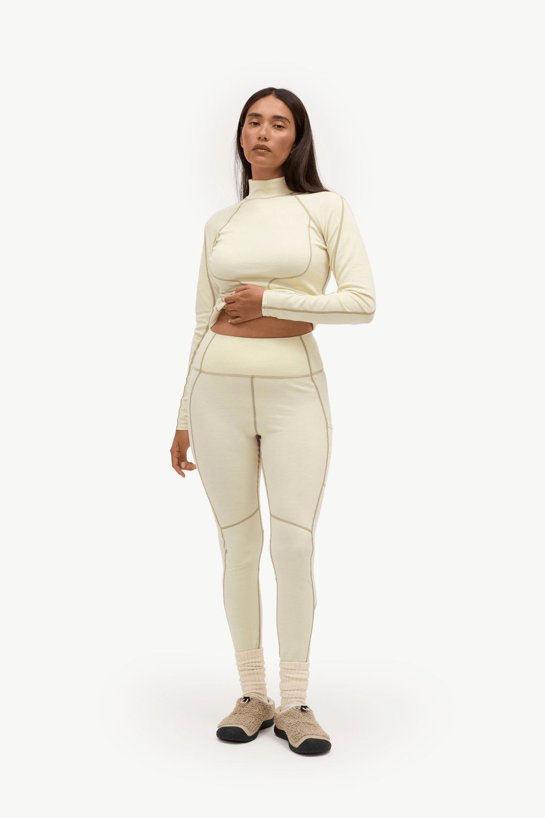 A woman is shown wearing a matching cream-colored activewear set consisting of a long-sleeve top and high-waisted leggings with contrast stitching, paired with beige socks and black shoes, against a plain white background.