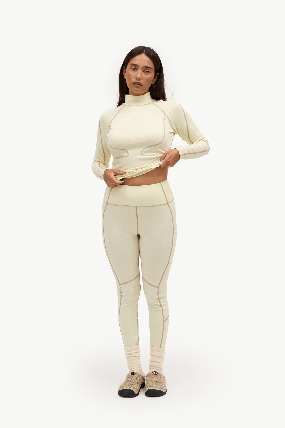 A woman modeling a matching cream-colored athletic set consisting of a long-sleeve mock-neck crop top and high-waisted leggings, paired with beige socks and tan shoes, against a plain white studio background.