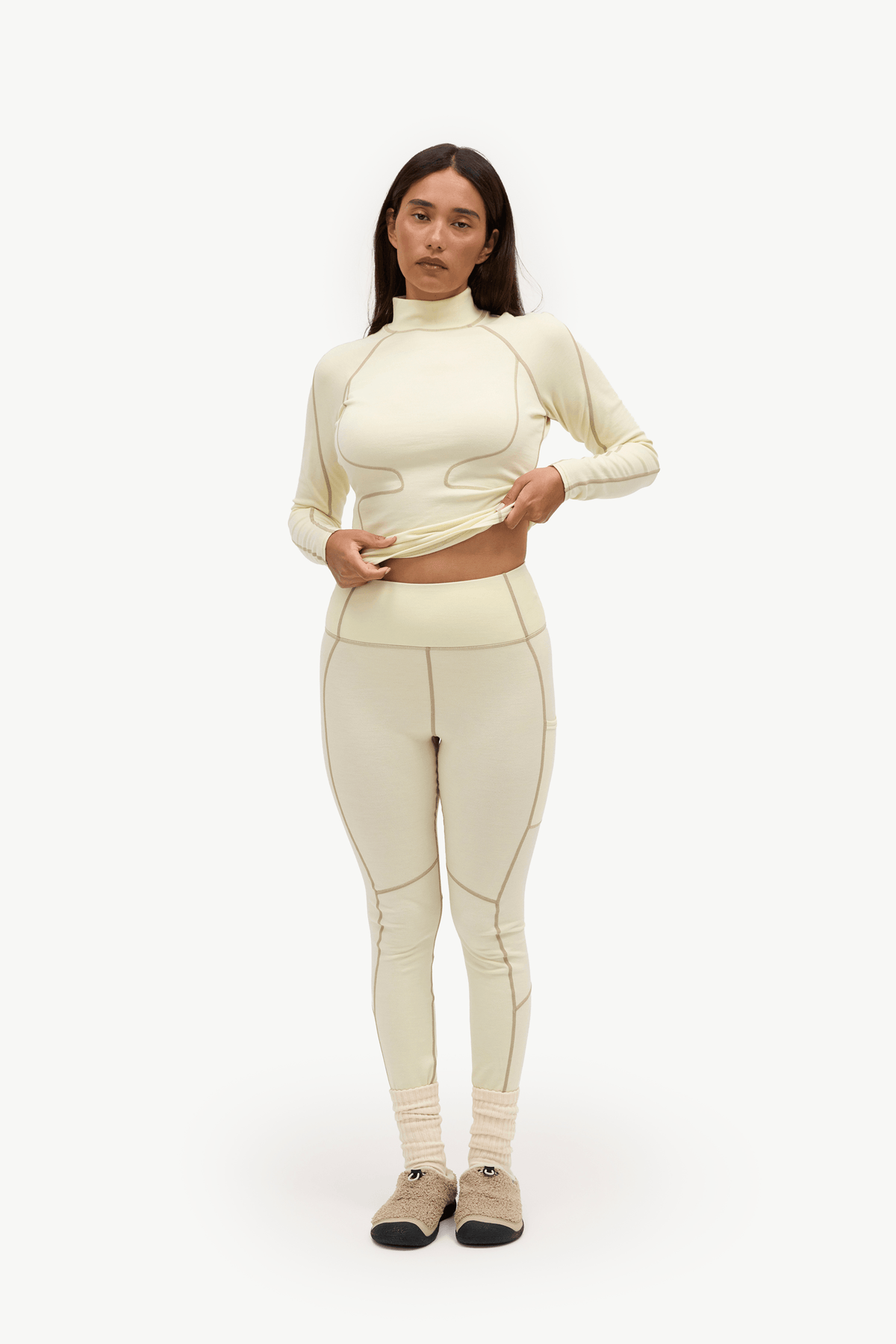 A woman modeling a matching cream-colored athletic set consisting of a long-sleeve mock-neck crop top and high-waisted leggings, paired with beige socks and tan shoes, against a plain white studio background.