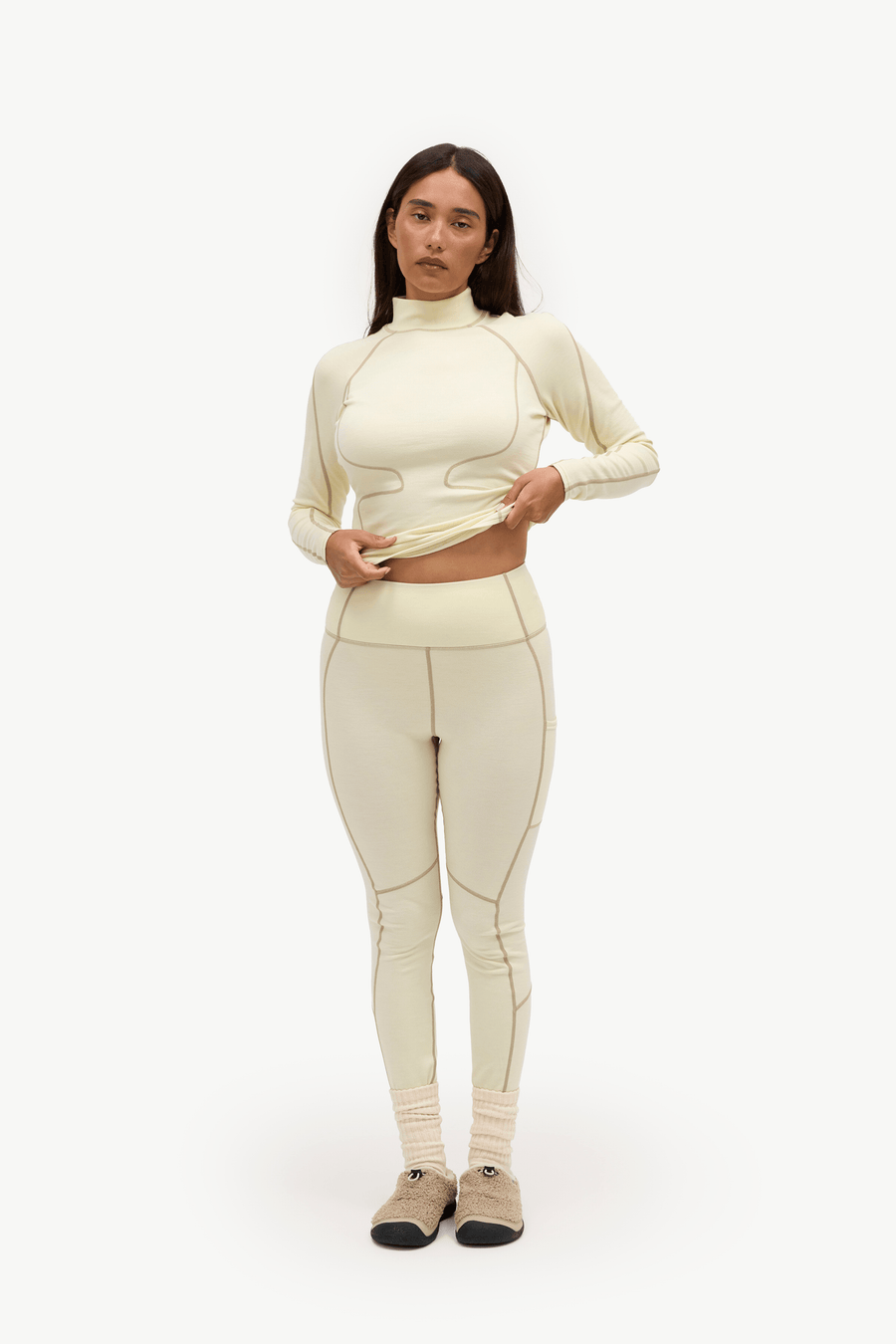 A woman modeling a matching cream-colored athletic set consisting of a long-sleeve mock-neck crop top and high-waisted leggings, paired with beige socks and tan shoes, against a plain white studio background.