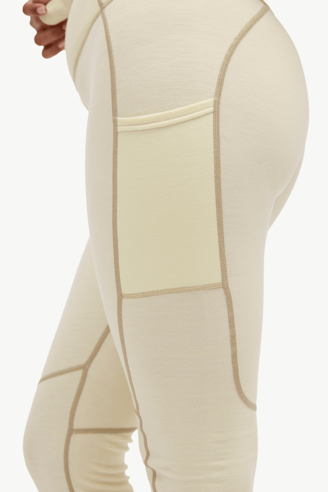 Close-up side view of a person wearing high-waisted leggings in a warm off-white/cream color with contrasting tan seam lines and a side pocket, against a plain light background.
