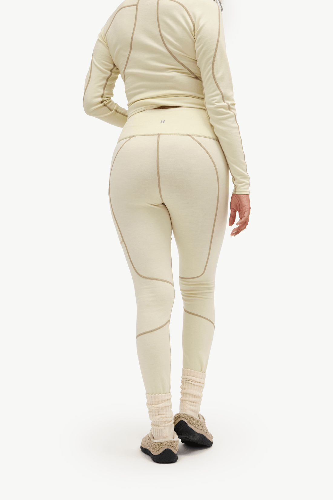 A person shown from the back wearing a matching cream athletic set with a long-sleeve top and high-waisted leggings, paired with beige socks and black shoes, against a plain white studio background.