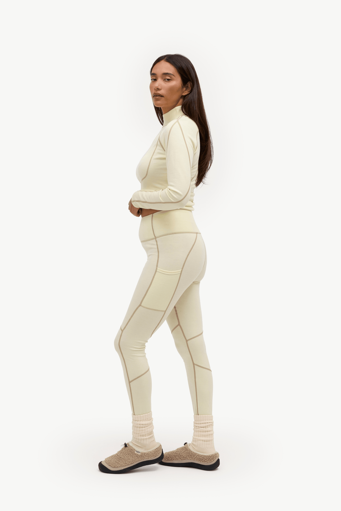 A woman modeling a matching cream-colored activewear set consisting of a mock-neck long-sleeve top and high-waisted leggings with geometric panel details, paired with beige knit socks and beige slip-on shoes, against a plain white studio background.