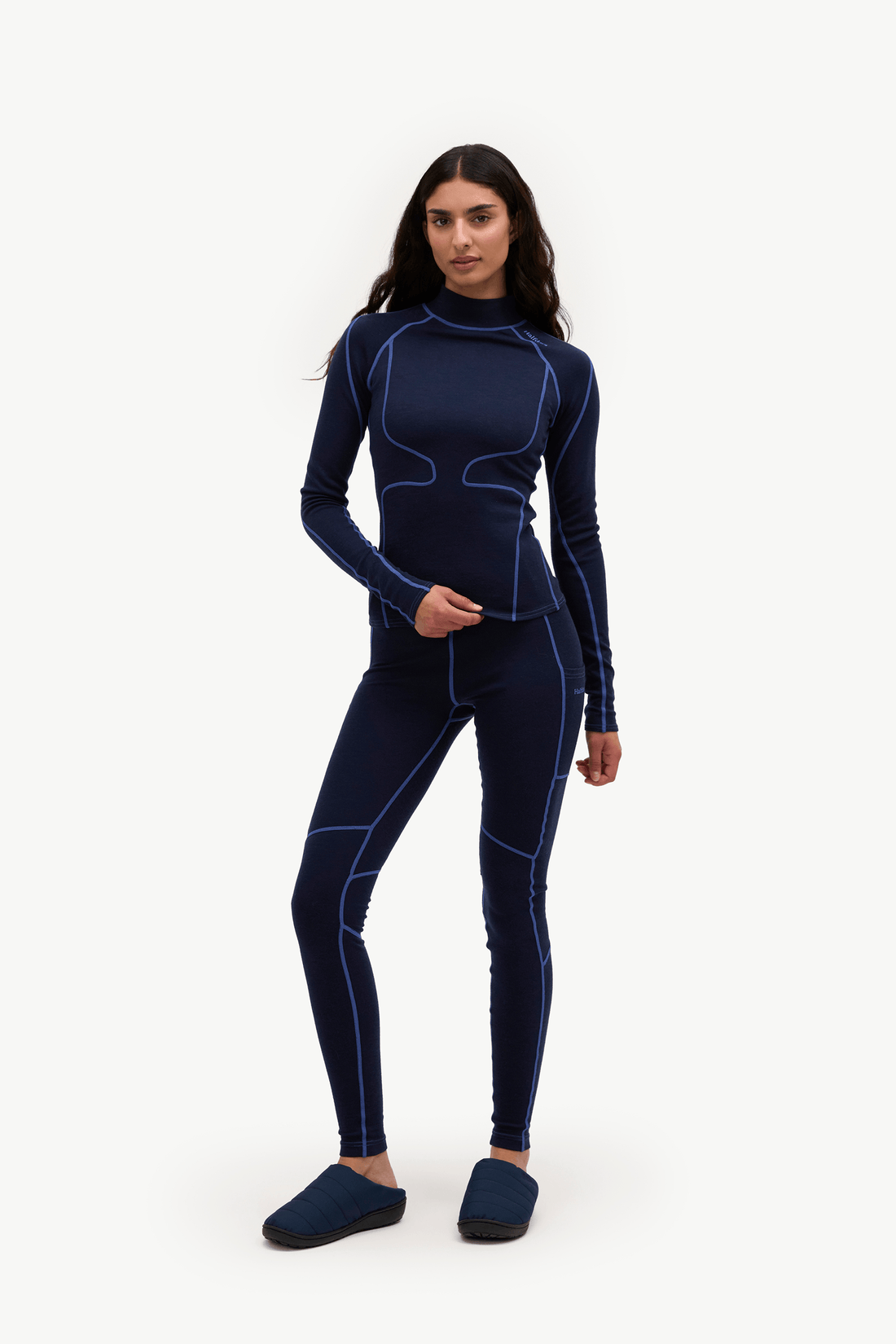 A woman modeling a dark navy blue two-piece wetsuit with contrasting light blue seam lines, paired with matching dark blue slippers, against a plain white studio background.