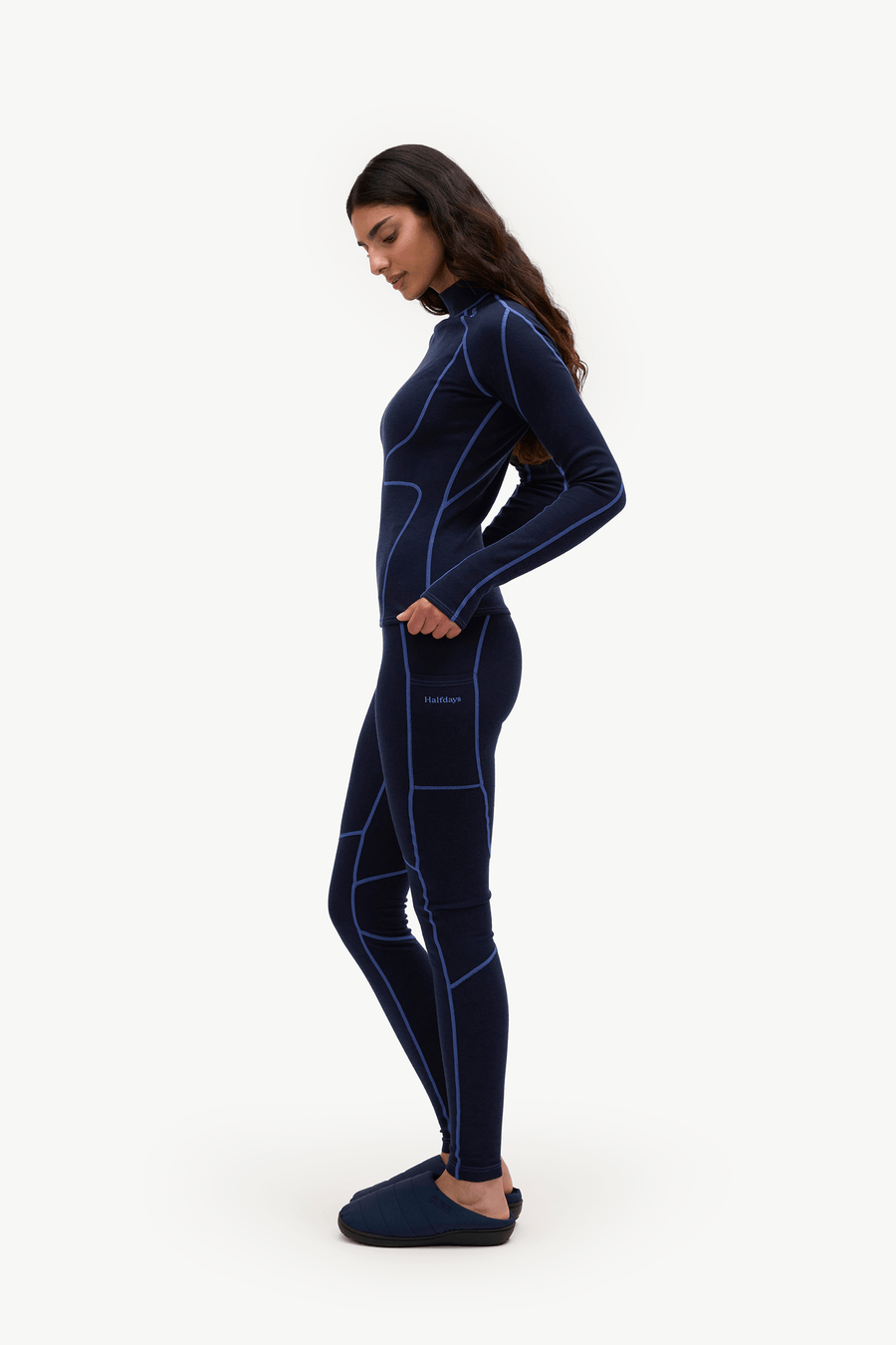 A woman wearing a navy blue full-body wetsuit with bright blue seam detailing, standing in profile against a plain white studio background.