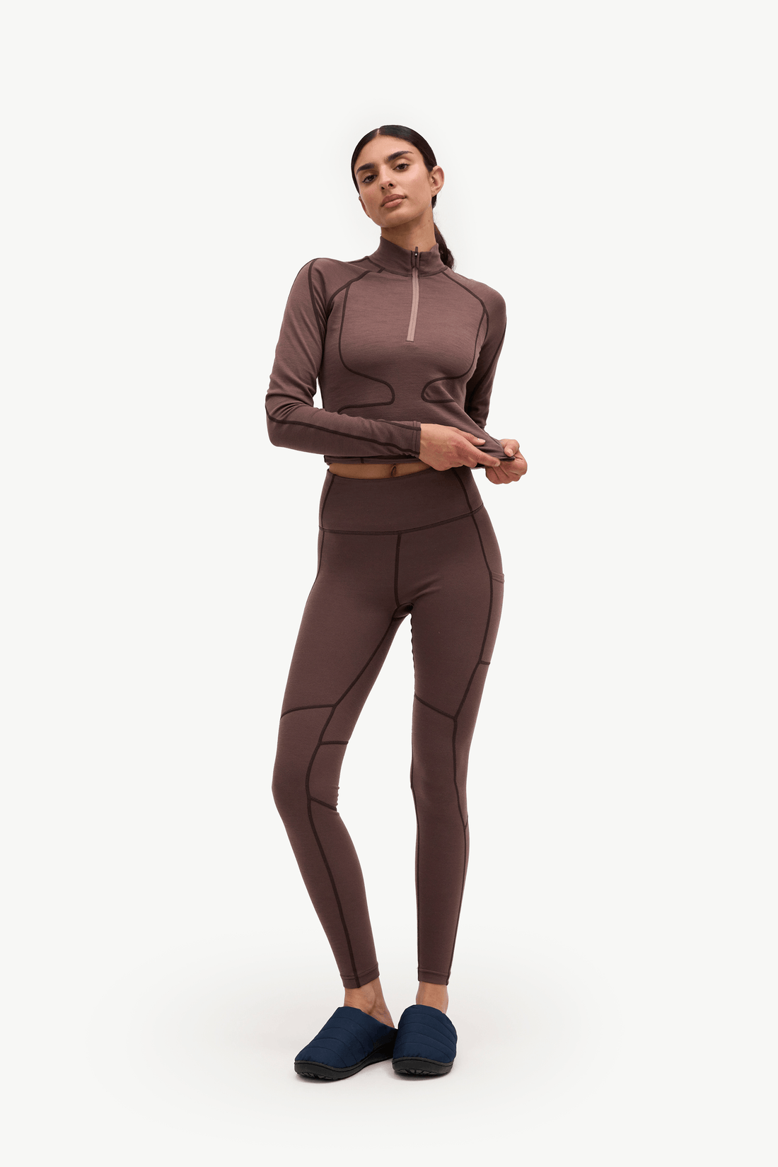 A woman modeling a matching athletic set consisting of a long-sleeve quarter-zip top and high-waisted leggings in a dark brown color, paired with dark blue slip-on shoes, against a plain white studio background.