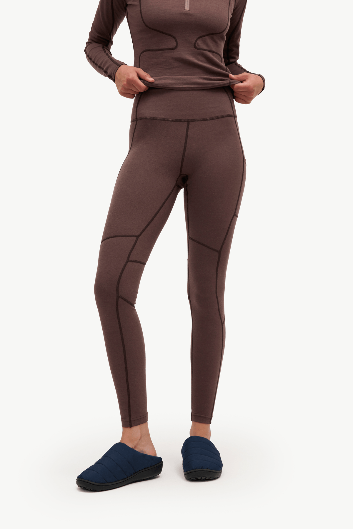 A model wearing a brown long-sleeve athletic top and matching high-waisted leggings with dark contrast stitching, paired with dark blue slip-on slippers, against a plain white background.