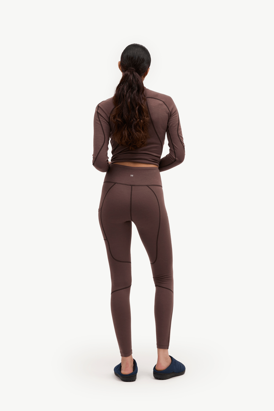 A woman viewed from behind wearing a matching brown activewear set consisting of a long-sleeve crop top and high-waisted leggings, paired with dark blue slippers, against a plain white background.