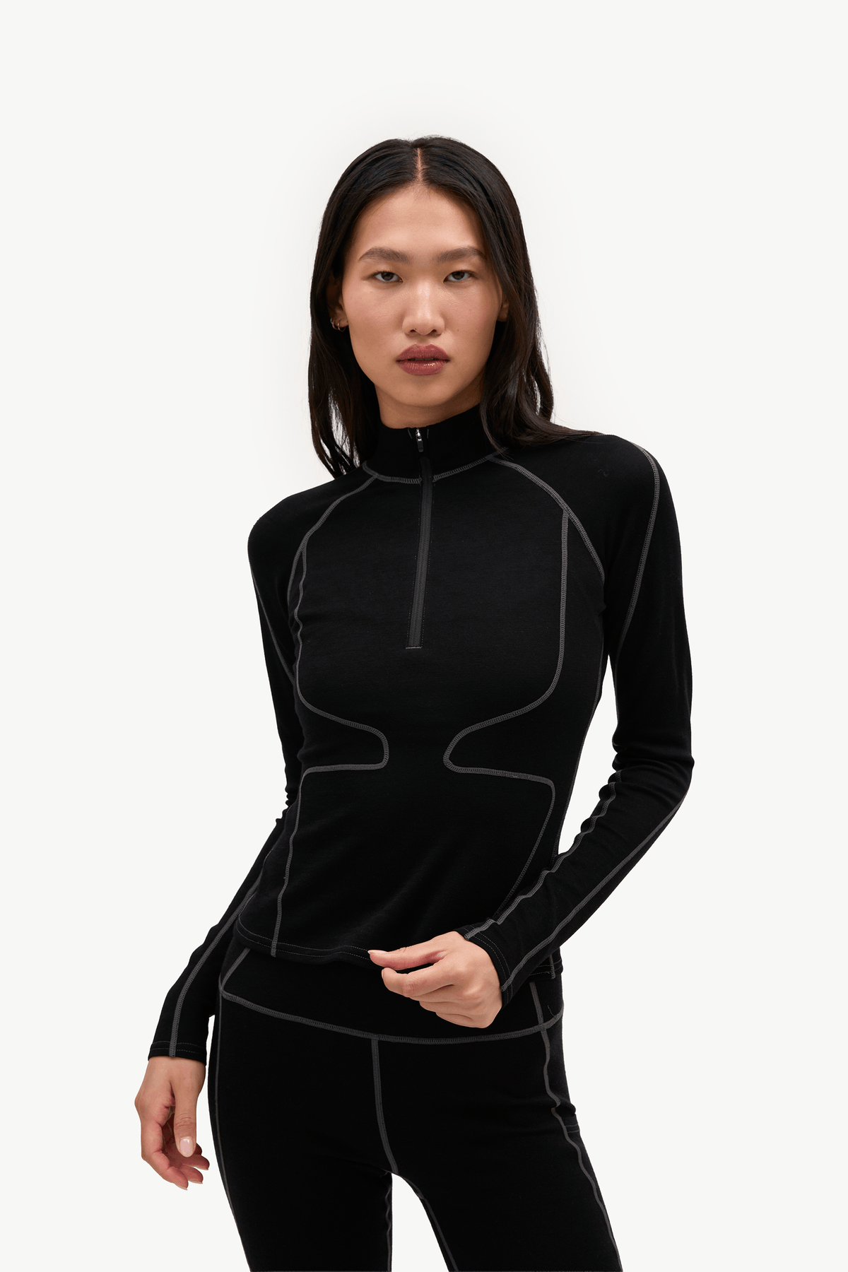 A woman modeling a black long-sleeve athletic quarter-zip top with contrasting light gray seam lines, paired with black leggings, against a plain white studio background.