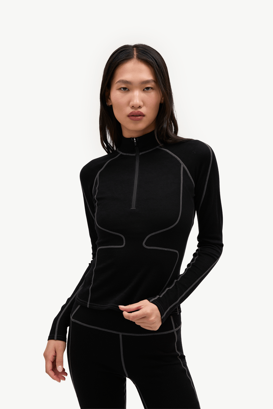 A woman modeling a black long-sleeve athletic quarter-zip top with contrasting light gray seam lines, paired with black leggings, against a plain white studio background.