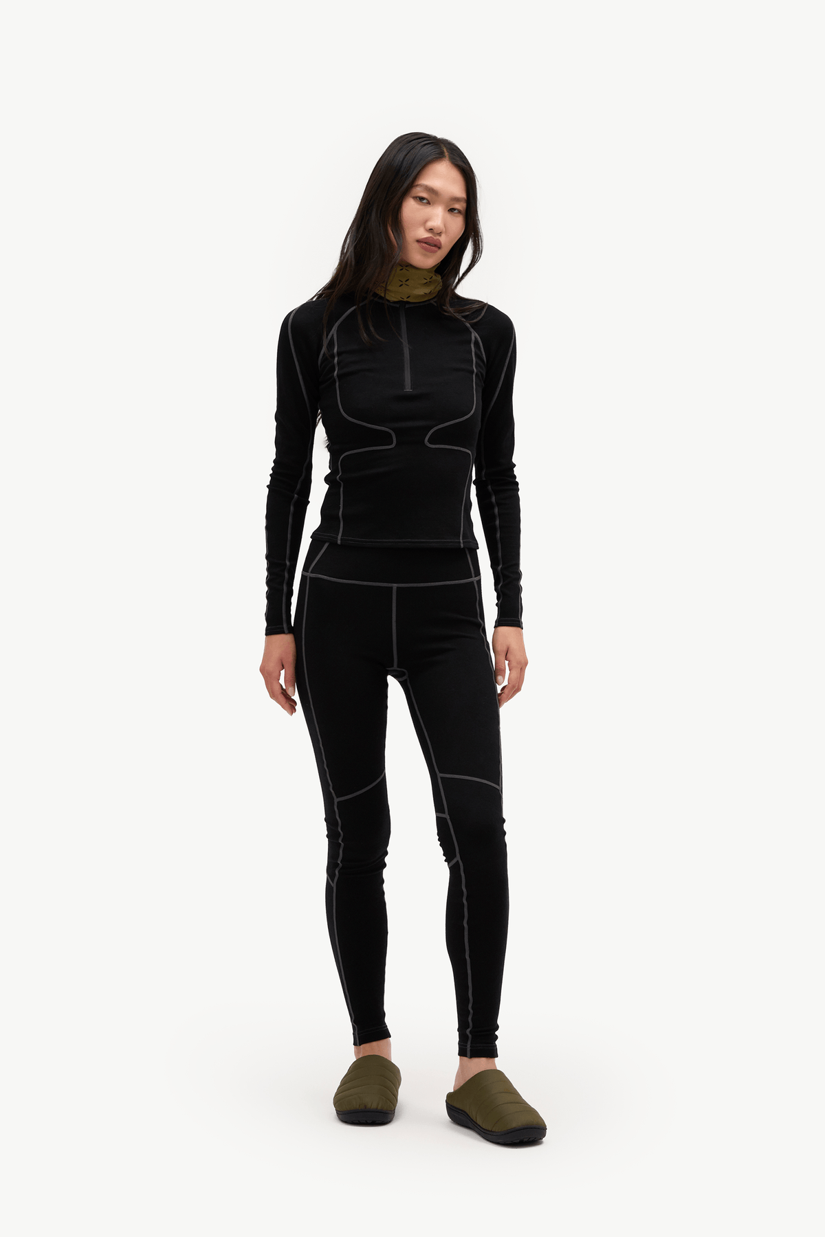 A woman modeling a black long-sleeve athletic top and matching black leggings with subtle contrast stitching, paired with olive green slip-on shoes, against a plain white studio background.
