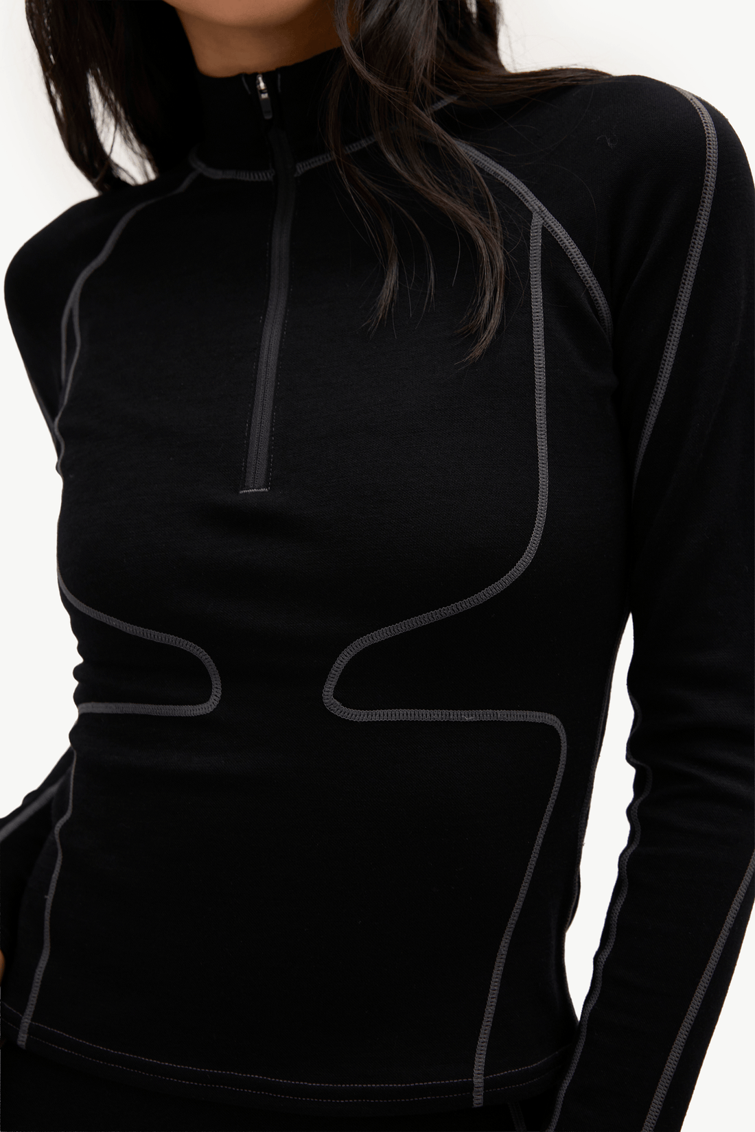 A close-up of a woman wearing a black long-sleeve athletic quarter-zip top with contrasting gray seam piping, shown against a plain light background.