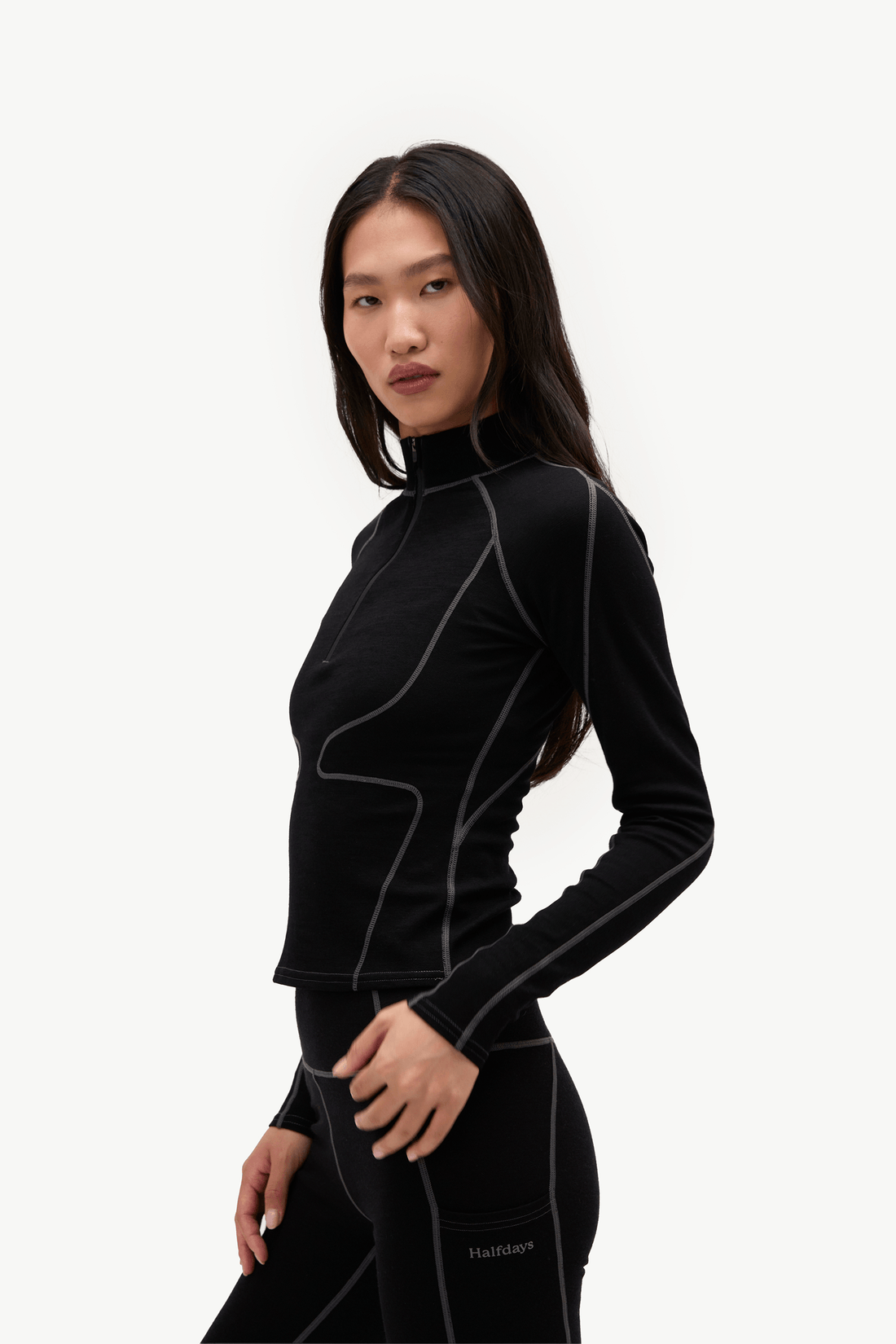 A woman modeling a black long-sleeve athletic top and matching leggings with light gray contrast stitching, photographed against a clean white studio background.