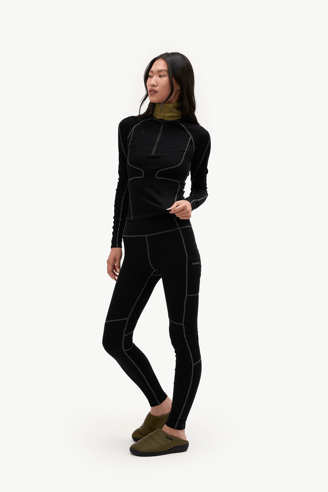 A woman modeling a black long-sleeve athletic top and matching black leggings with white contrast stitching, paired with olive green slip-on shoes, against a plain white background.