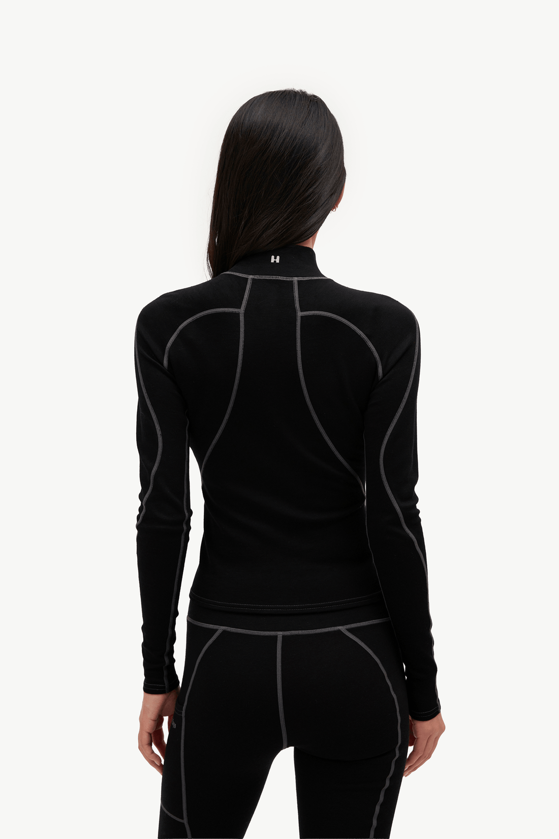 A woman viewed from the back wearing a black long-sleeve athletic top and matching black leggings with contrasting gray seam detailing, against a plain light gray studio background.