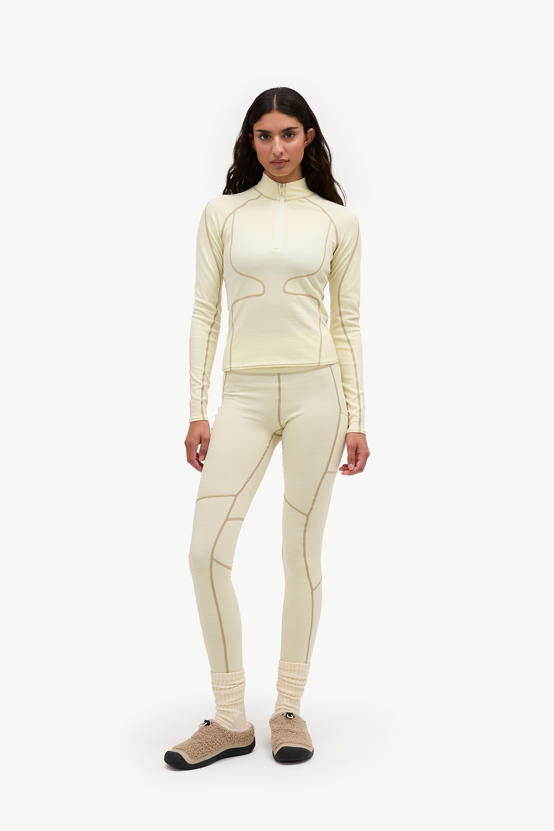 A model stands against a light gray studio background wearing a matching cream-colored athletic set consisting of a long-sleeve mock-neck top and high-waisted leggings with contrasting tan seam detailing, paired with beige knit socks and tan sneakers.