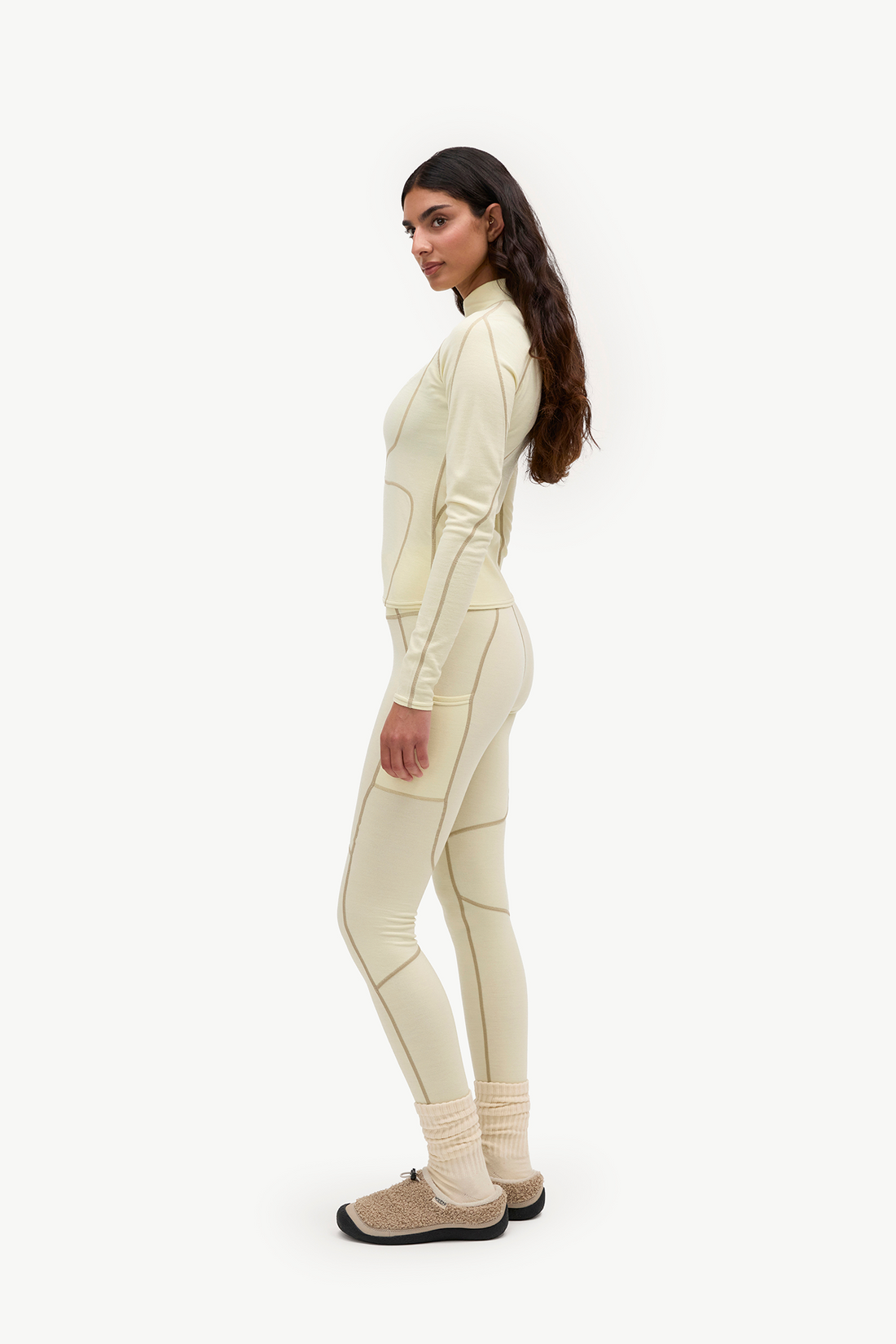 A woman stands in profile wearing a matching cream-colored athletic tracksuit with contouring panels and ribbed knit socks, paired with beige slip-on shoes, against a plain white studio background.