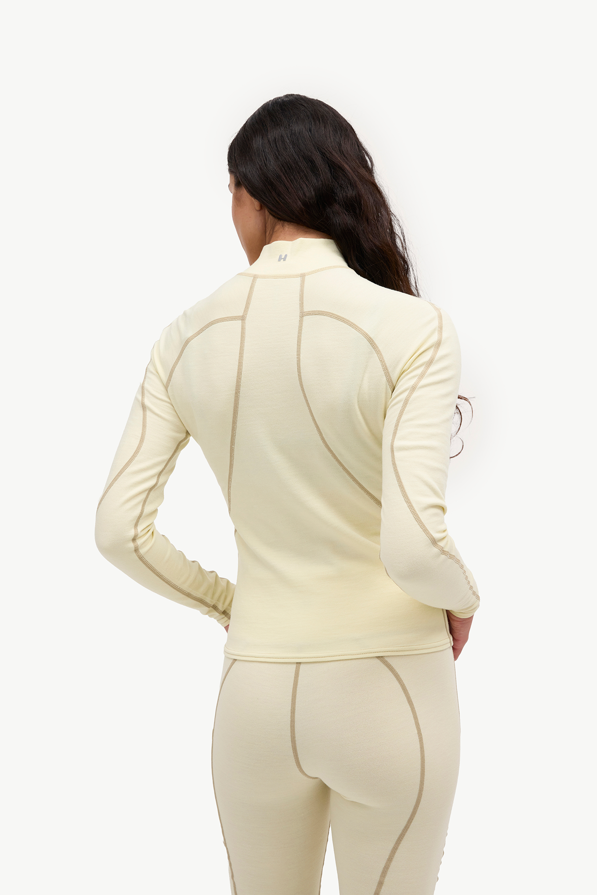 A woman shown from the back wearing a fitted long-sleeve athletic top and matching leggings in a light cream color with contrasting seam lines, against a plain white studio background.