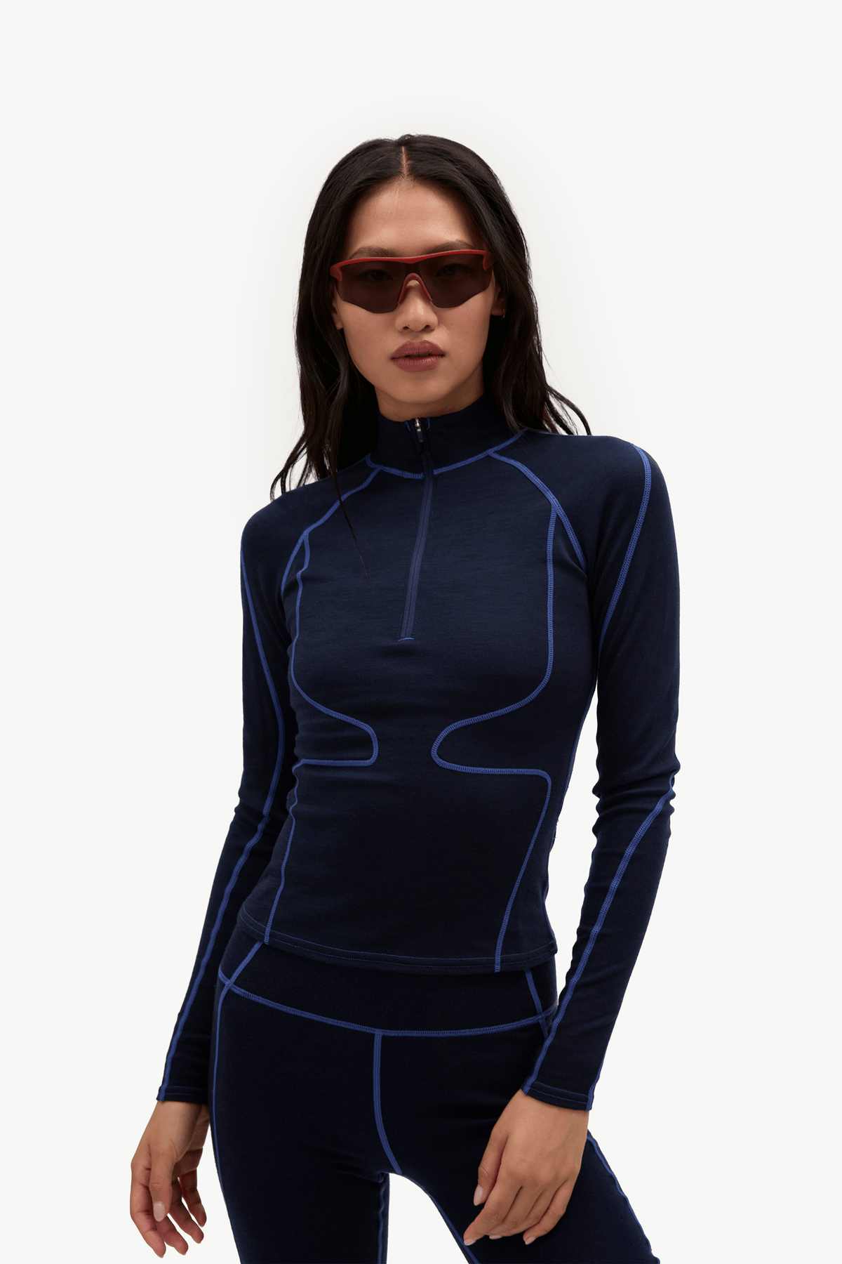 A woman modeling a dark navy long-sleeve athletic top with bright blue contrast stitching and a half-zip neckline, paired with matching dark leggings, against a plain white studio background.