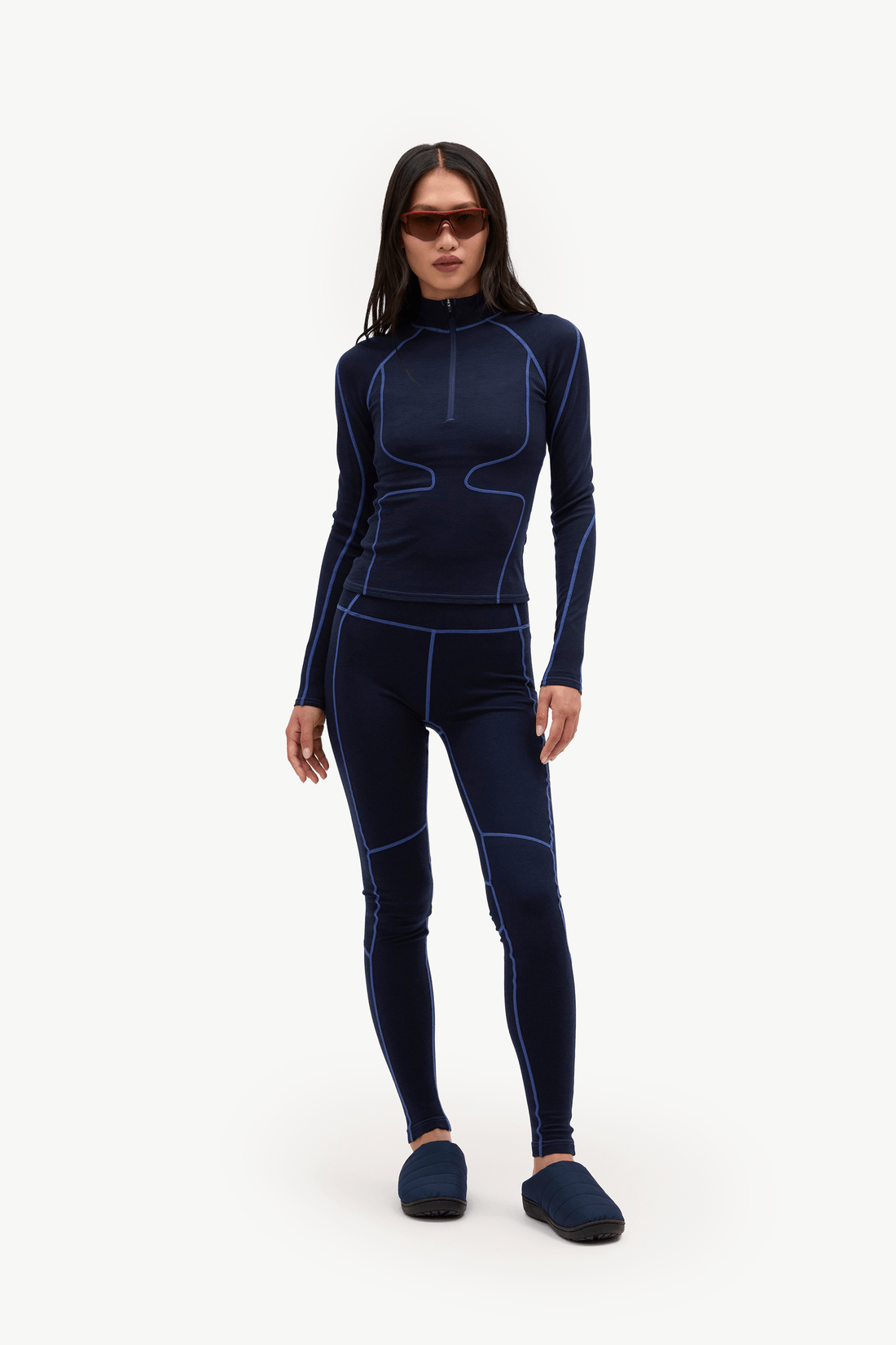 A woman modeling a dark navy blue full-body wetsuit with contrasting bright blue seam lines, paired with dark blue slippers, against a plain white studio background.