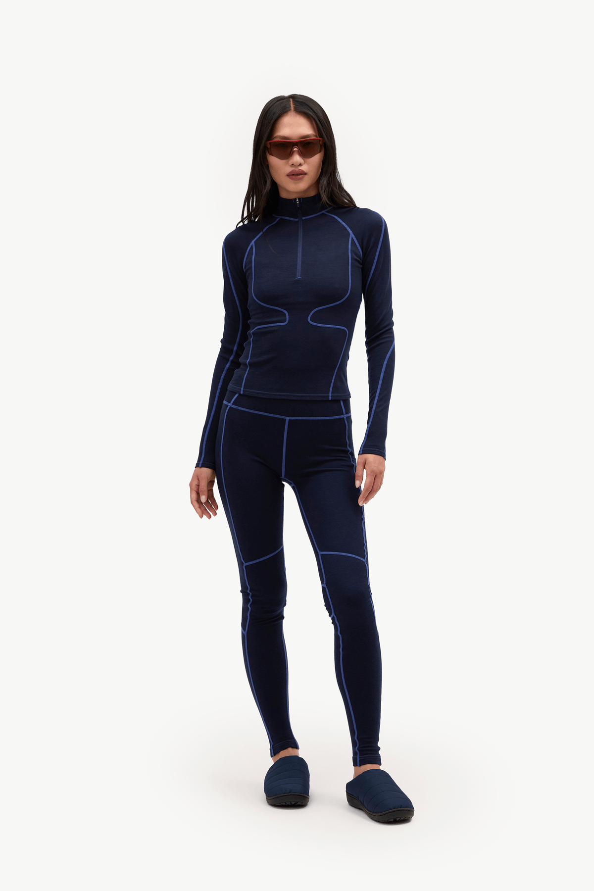 A woman modeling a dark navy blue full-body wetsuit with contrasting bright blue seam lines, paired with dark blue slippers, against a plain white studio background.