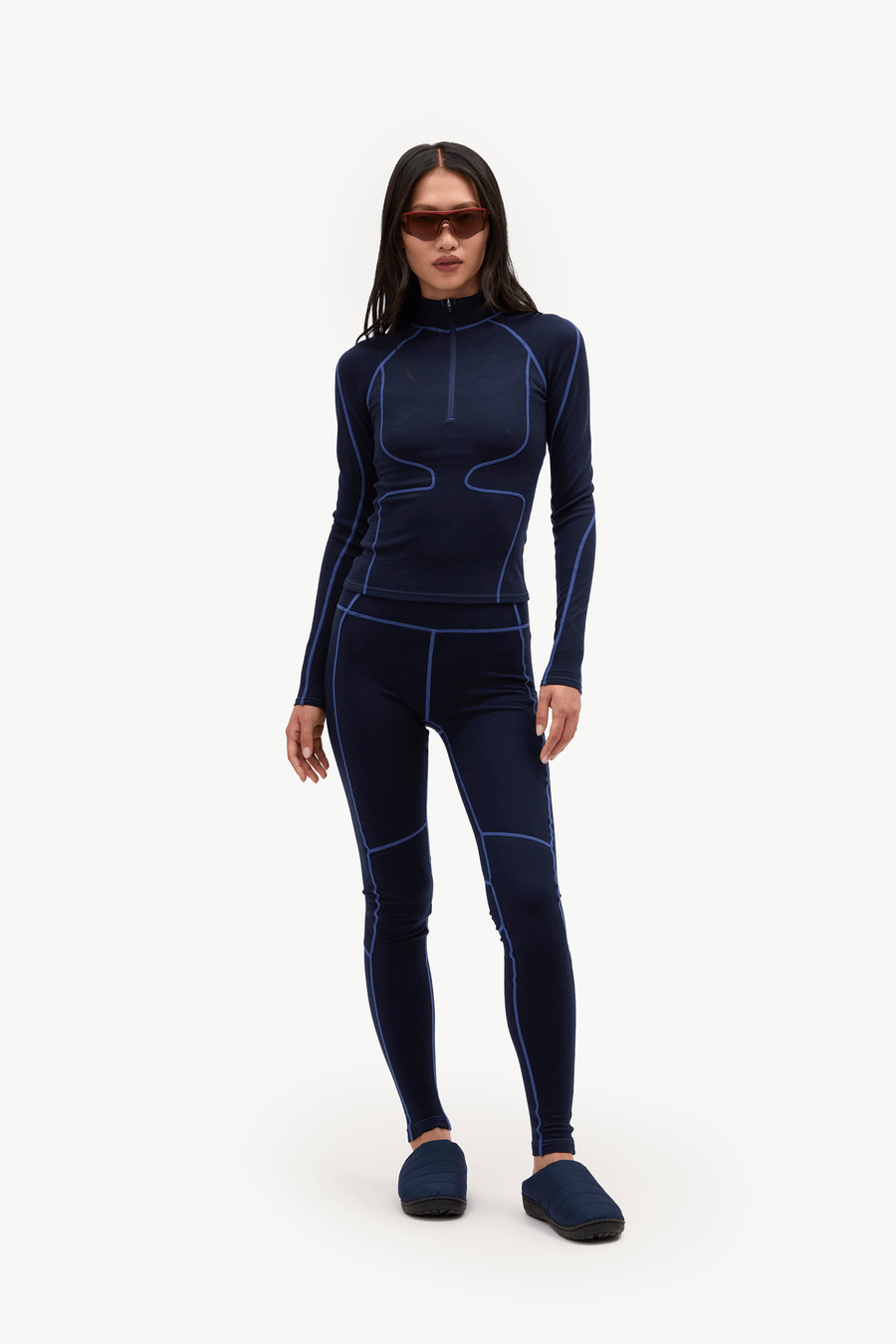 A woman modeling a dark navy blue full-body wetsuit with contrasting bright blue seam lines, paired with dark blue slippers, against a plain white studio background.