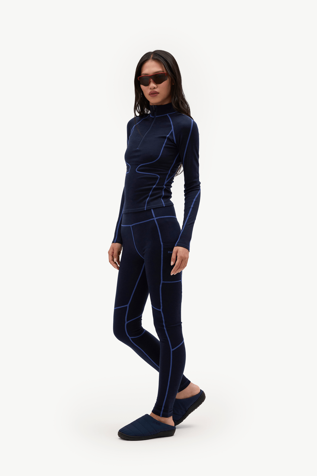 A woman modeling a dark navy blue two-piece athletic set consisting of a long-sleeve mock-neck top and matching leggings with bright blue contrast stitching, paired with dark navy slip-on shoes, against a plain white studio background.