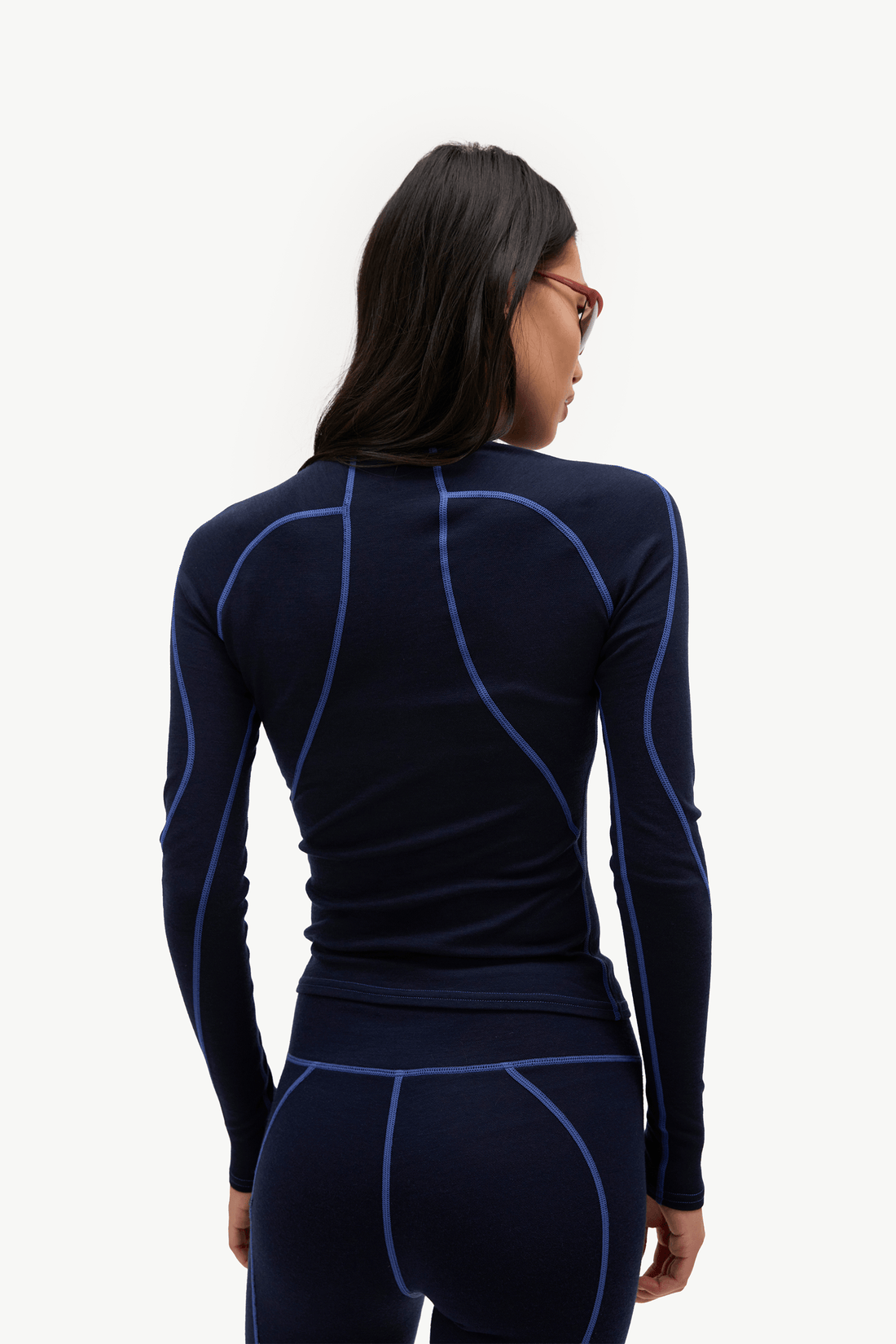 A woman shown from the back wearing a dark navy long-sleeve athletic compression top with bright blue seam detailing, paired with matching navy leggings, against a plain white background.
