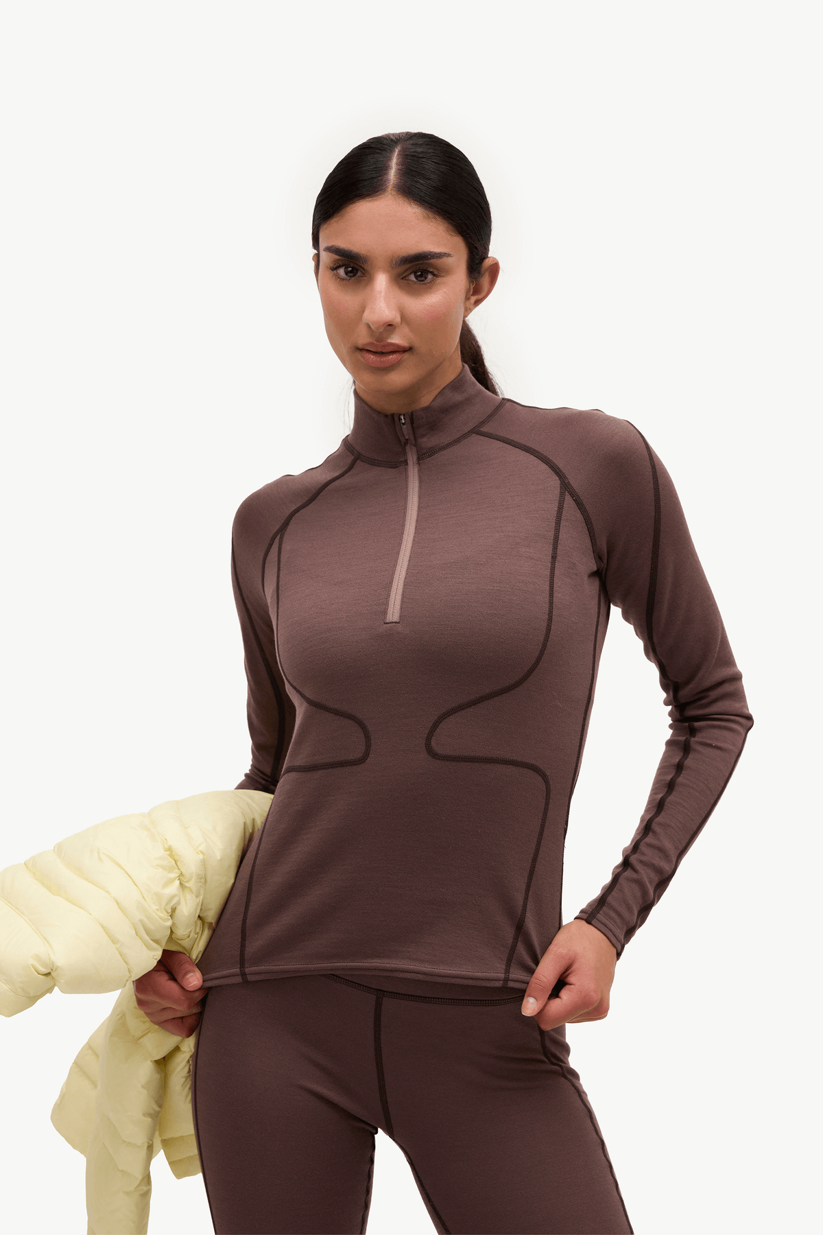 A woman modeling a brown long-sleeve athletic half-zip top and matching leggings, holding a light yellow puffer jacket against a plain white studio background.
