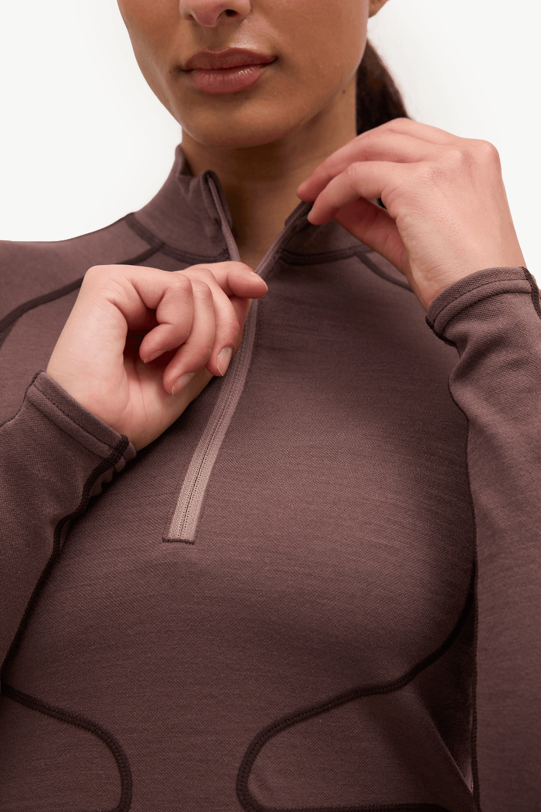 Close-up of a woman wearing a brown long-sleeve quarter-zip athletic top, adjusting the zipper with both hands against a plain white background.