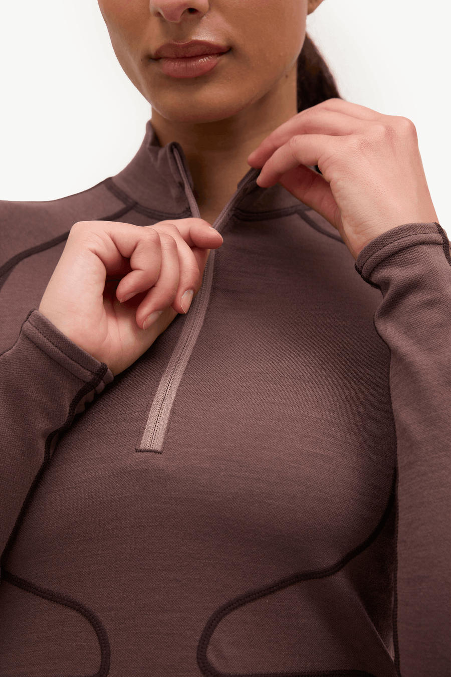 Close-up of a woman wearing a brown long-sleeve quarter-zip athletic top, adjusting the zipper with both hands against a plain white background.