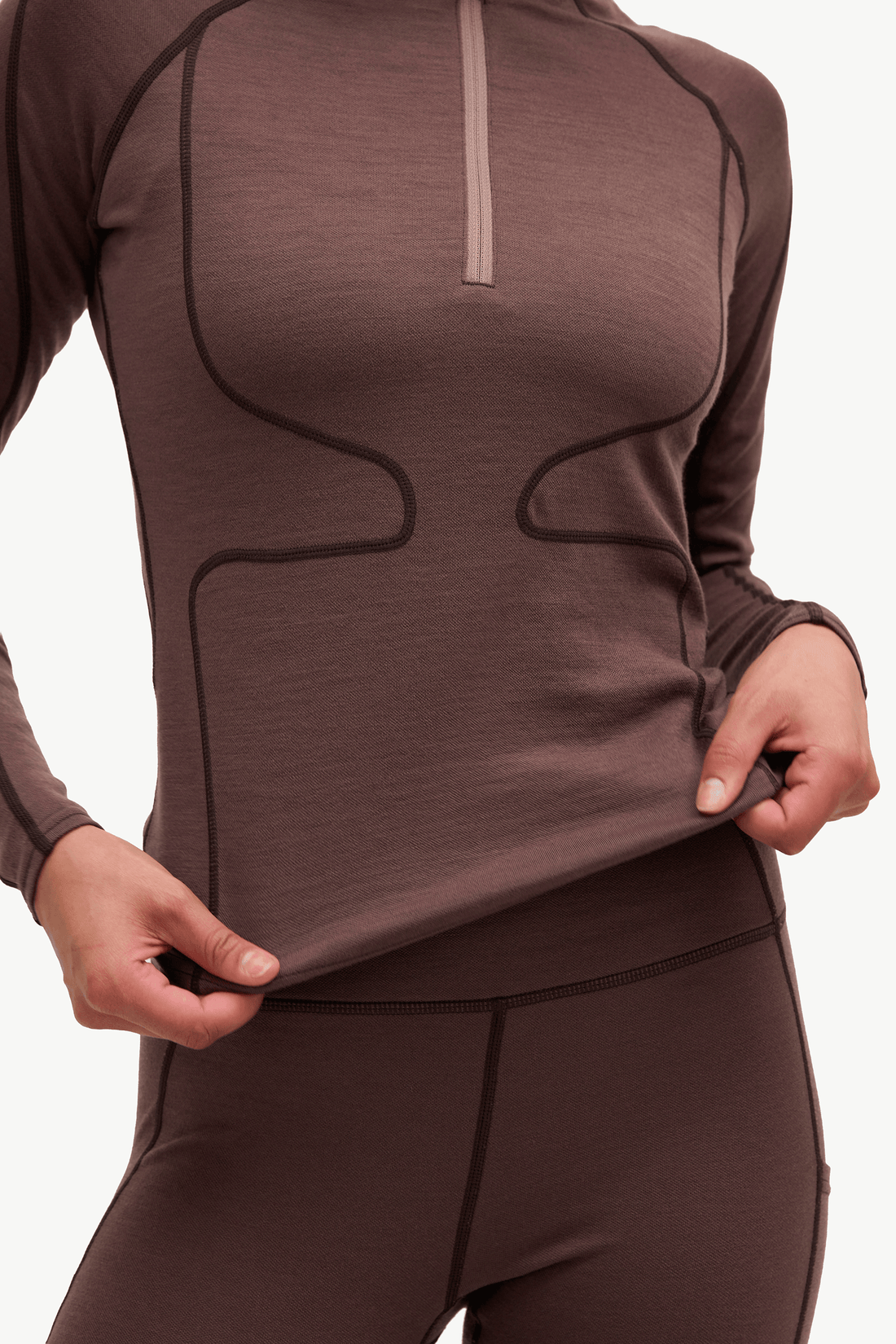 A close-up of a person wearing a fitted long-sleeve athletic top and matching leggings in a dark taupe-brown color, with contrast darker piping and a light zipper placket.