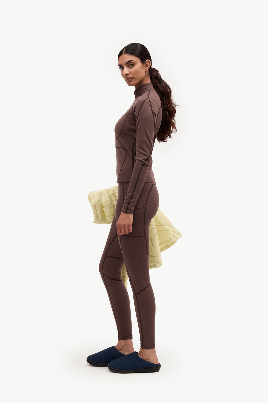 A woman stands in profile wearing a fitted brown long-sleeve top and matching leggings, with dark navy slip-on shoes, against a plain light background. She is holding a pale yellow garment behind her back.