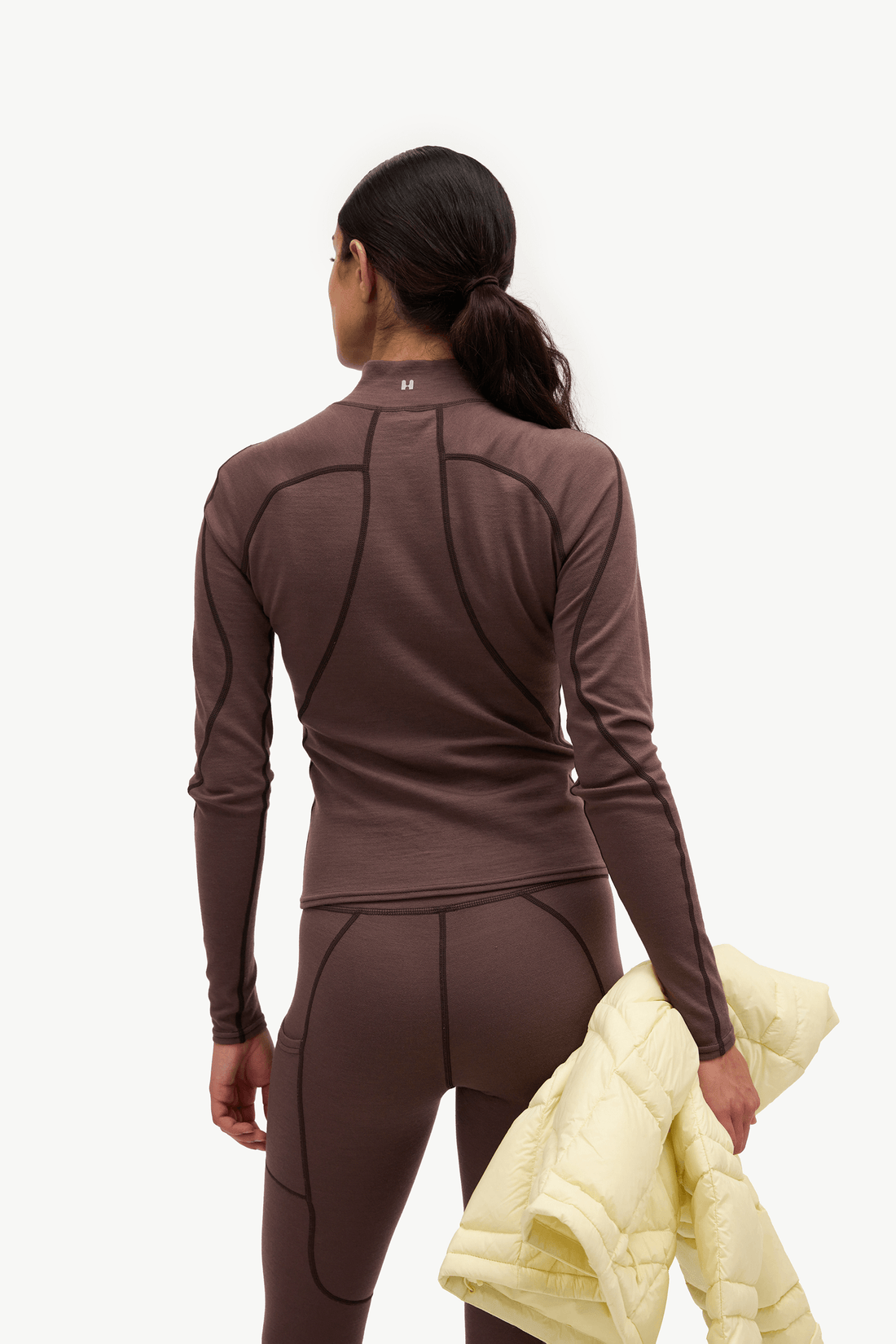 A woman shown from the back wearing a fitted long-sleeve athletic top and matching leggings in a dark brown color, holding a pale yellow puffer jacket against a plain white studio background.