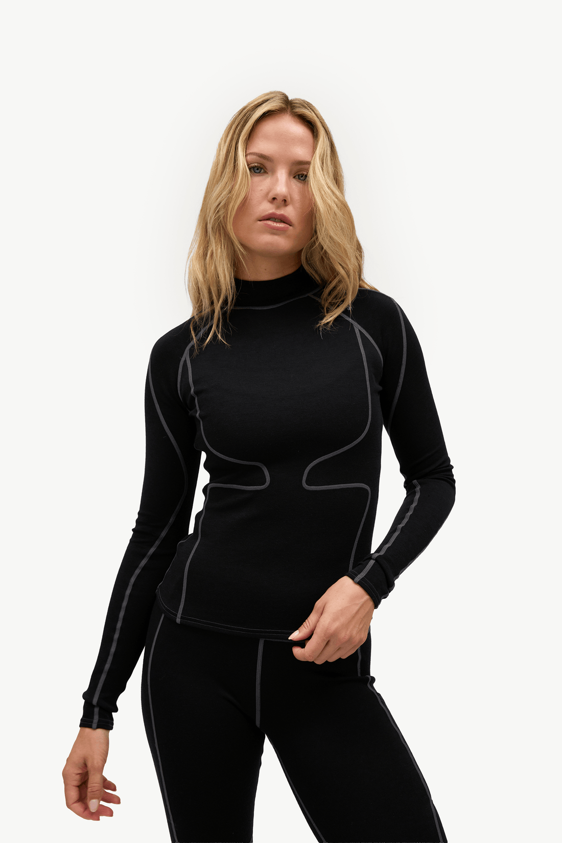 A blonde woman modeling a black long-sleeve athletic top with contrasting gray piping details, paired with black leggings, against a plain light gray background.