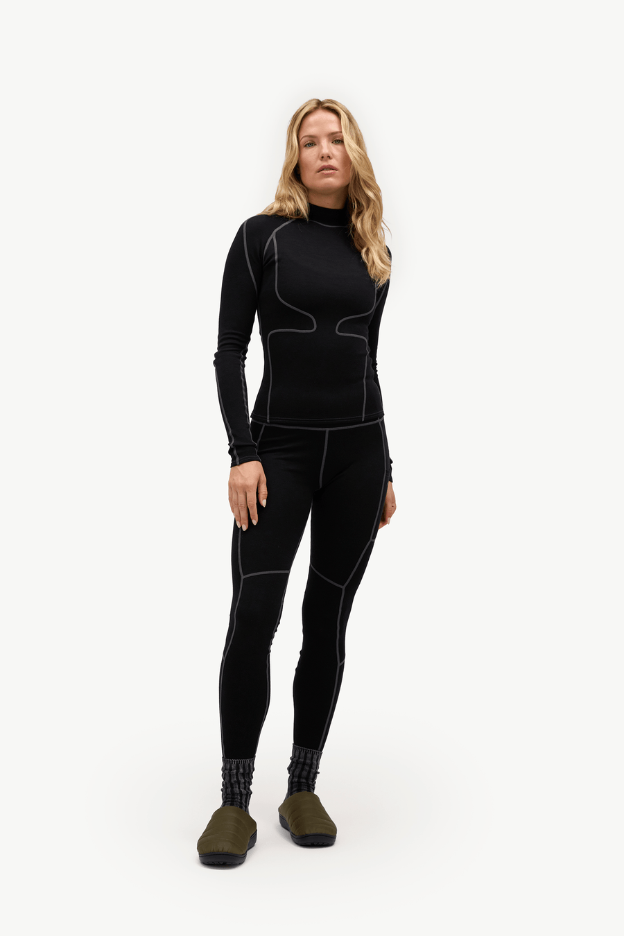 A woman modeling a black long-sleeve athletic wetsuit with white contrast stitching, paired with black leggings and olive green slippers, against a plain white studio background.